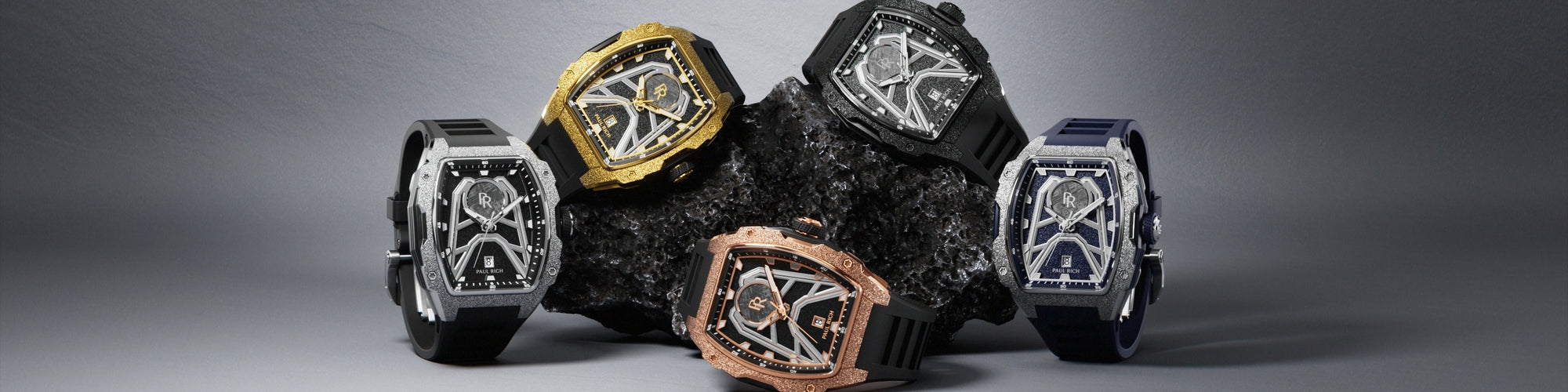 Five Paul Rich tonneau watches in silver, gold, rose gold, black, and blue displayed around a black volcanic rock on a gray backdrop. Five Paul Rich tonneau watches in silver, gold, rose gold, black, and blue displayed around a black volcanic rock on a gray backdrop.