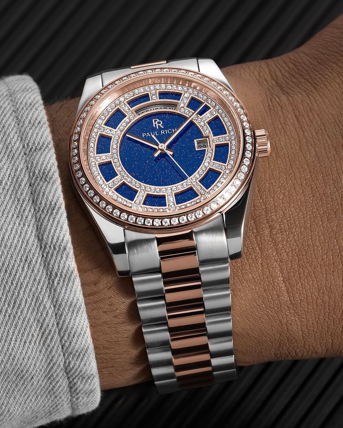 Rose gold and silver two-tone watch with a blue aventurine dial, pavé bezel, and day–date display shown on a wrist with a brushed metal bracelet. Rose gold and silver two-tone watch with a blue aventurine dial, pavé bezel, and day–date display shown on a wrist with a brushed metal bracelet.