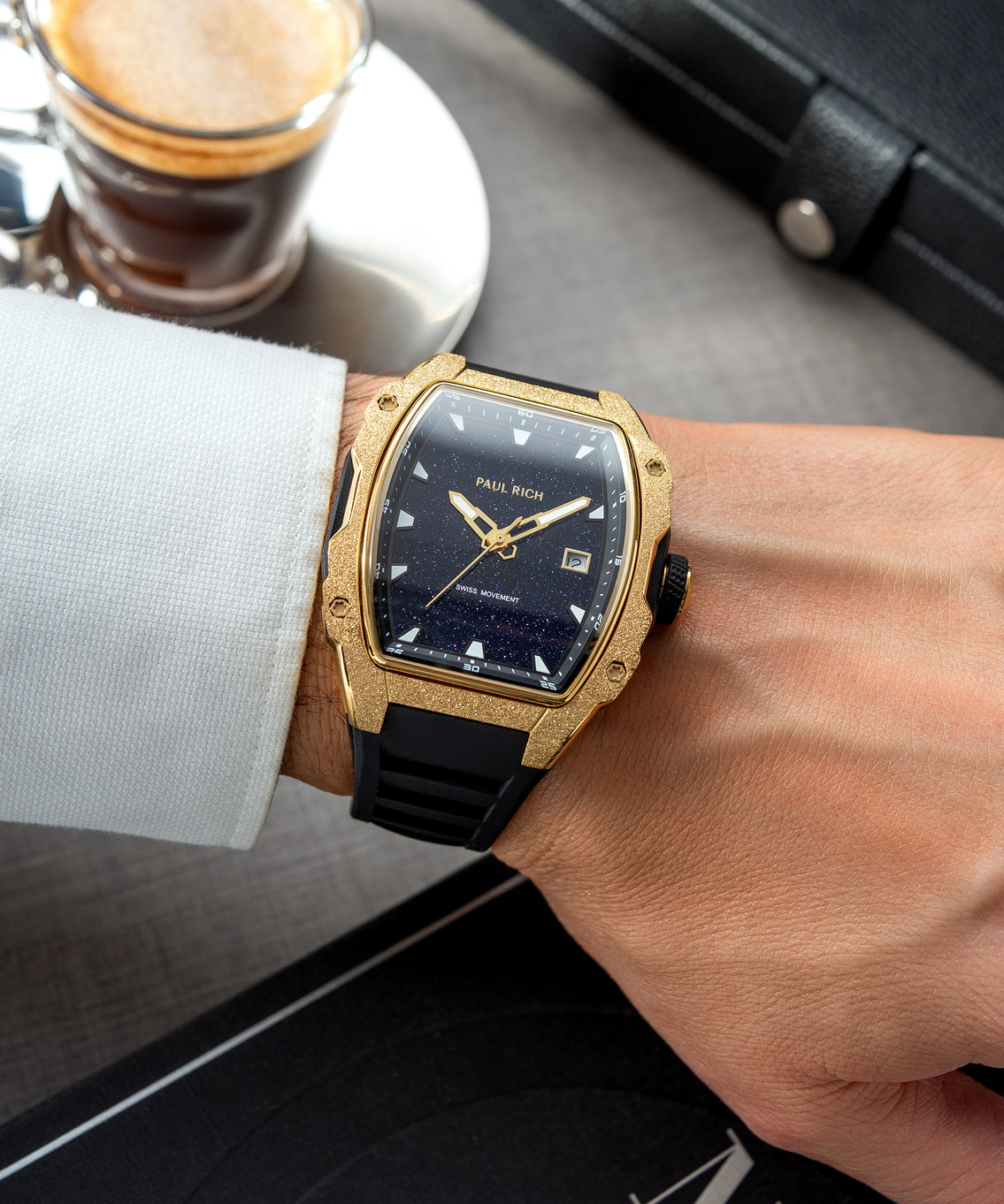 Tonneau-shaped Paul Rich watch with blue aventurine dial and gold frosted bezel worn on wrist with white cuff, coffee in background. Tonneau-shaped Paul Rich watch with blue aventurine dial and gold frosted bezel worn on wrist with white cuff, coffee in background.