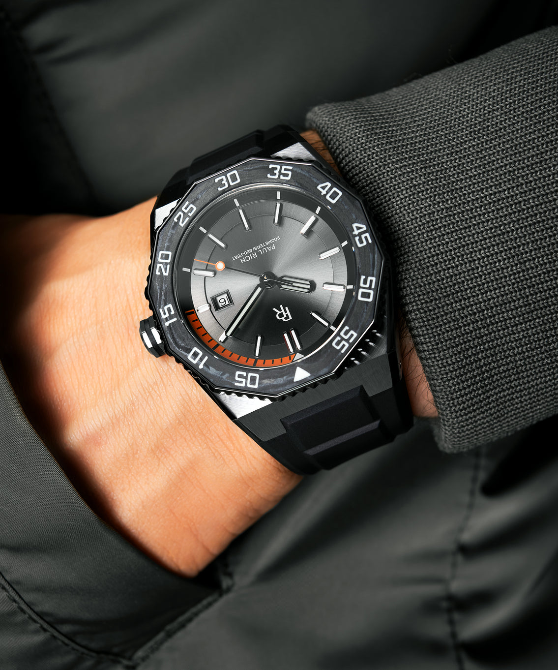 Paul Rich dive watch with silver sunray dial and orange dive timer worn on wrist, paired with a dark grey textured jacket. Paul Rich dive watch with silver sunray dial and orange dive timer worn on wrist, paired with a dark grey textured jacket.