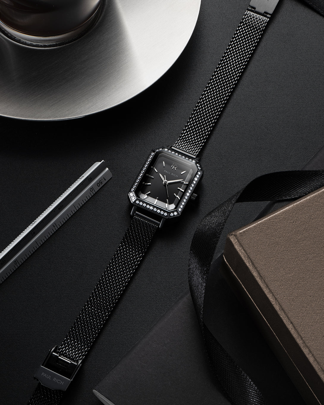 Black Paul Rich watch with black sunray dial, gem-set octagonal bezel, and black mesh strap, displayed flat on a dark surface alongside a cup of coffee, a ruler, a notebook, and a black ribbon. Black Paul Rich watch with black sunray dial, gem-set octagonal bezel, and black mesh strap, displayed flat on a dark surface alongside a cup of coffee, a ruler, a notebook, and a black ribbon.