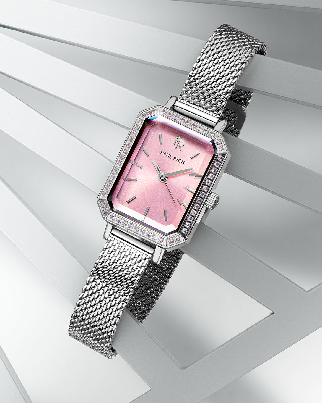 Silver-tone Paul Rich watch with pink sunray dial, pink gem-set octagonal bezel, and silver mesh strap, displayed diagonally across layered white geometric panels. Silver-tone Paul Rich watch with pink sunray dial, pink gem-set octagonal bezel, and silver mesh strap, displayed diagonally across layered white geometric panels.