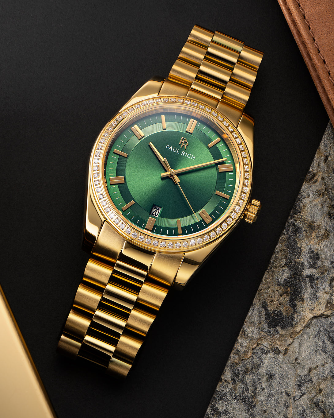 Gold-tone Paul Rich watch with green sunray dial, diamond-set bezel, and gold link bracelet, displayed on a flat lay surface with leather, stone, and matte black textures. Gold-tone Paul Rich watch with green sunray dial, diamond-set bezel, and gold link bracelet, displayed on a flat lay surface with leather, stone, and matte black textures.