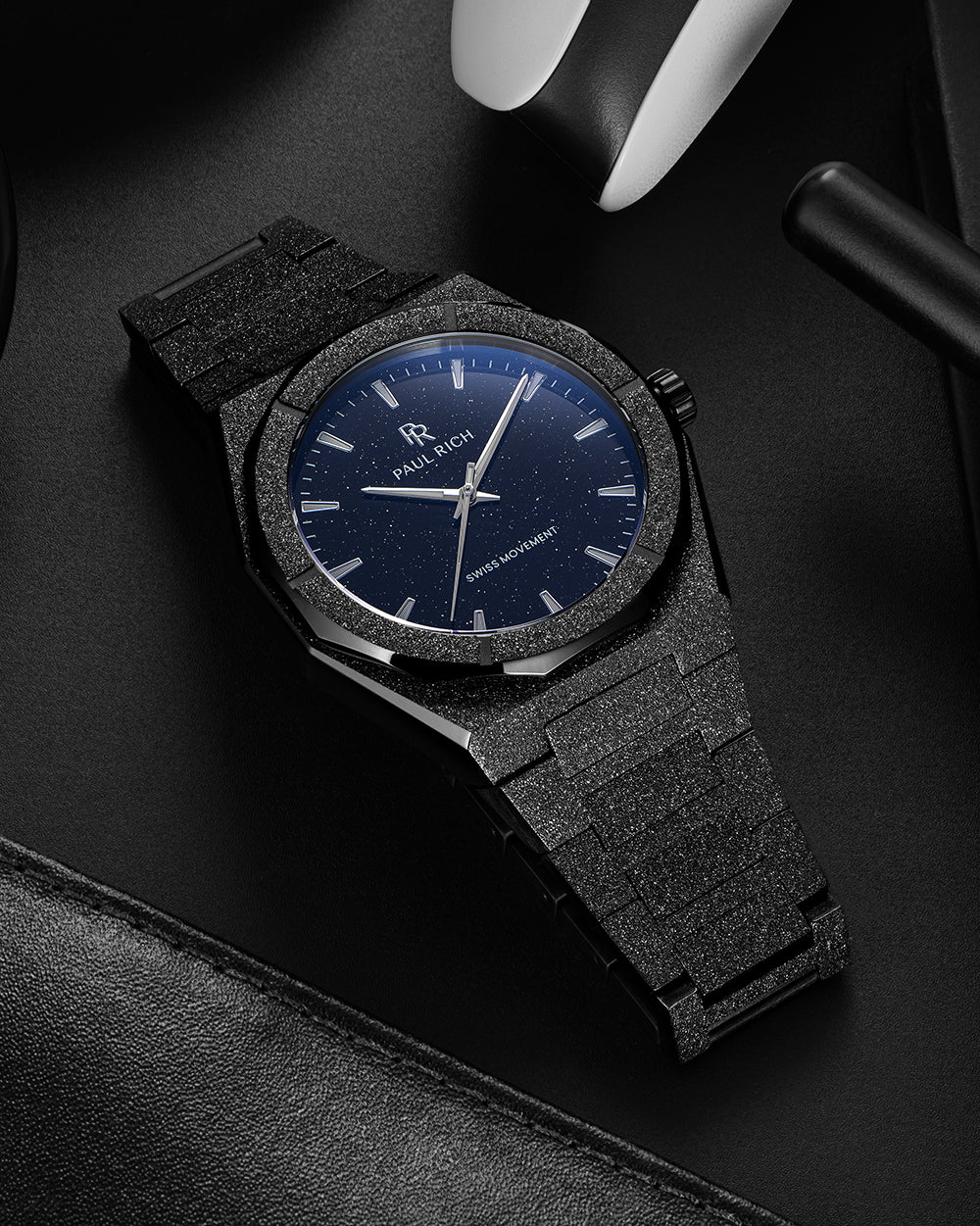 Black frosted Paul Rich watch with blue aventurine dial resting on a black leather surface near gaming gear and a headset. Black frosted Paul Rich watch with blue aventurine dial resting on a black leather surface near gaming gear and a headset.