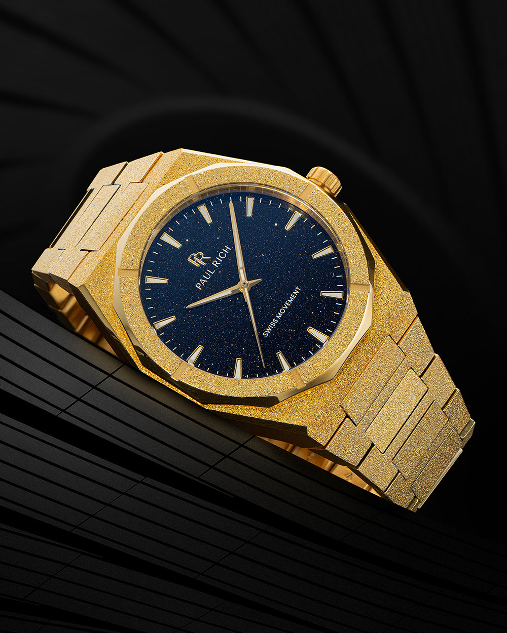 Gold frosted Paul Rich watch with blue aventurine dial and gold markers, resting on a sleek black geometric surface. Gold frosted Paul Rich watch with blue aventurine dial and gold markers, resting on a sleek black geometric surface.