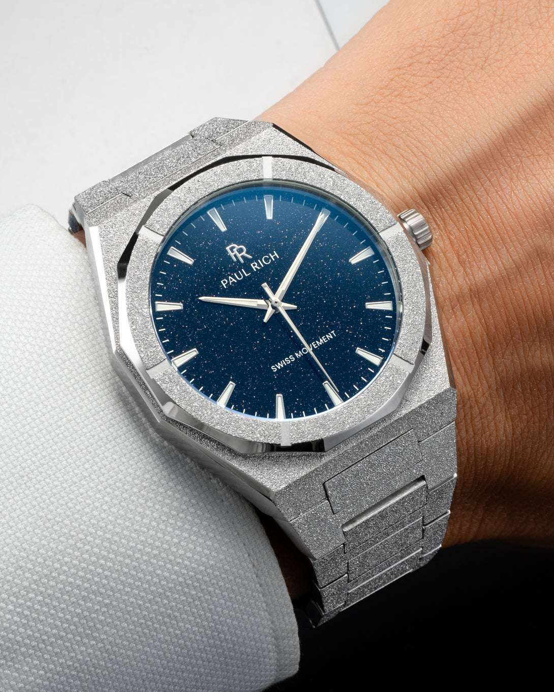 Silver frosted Paul Rich watch with blue aventurine dial worn on wrist, paired with a white textured dress shirt. Silver frosted Paul Rich watch with blue aventurine dial worn on wrist, paired with a white textured dress shirt.