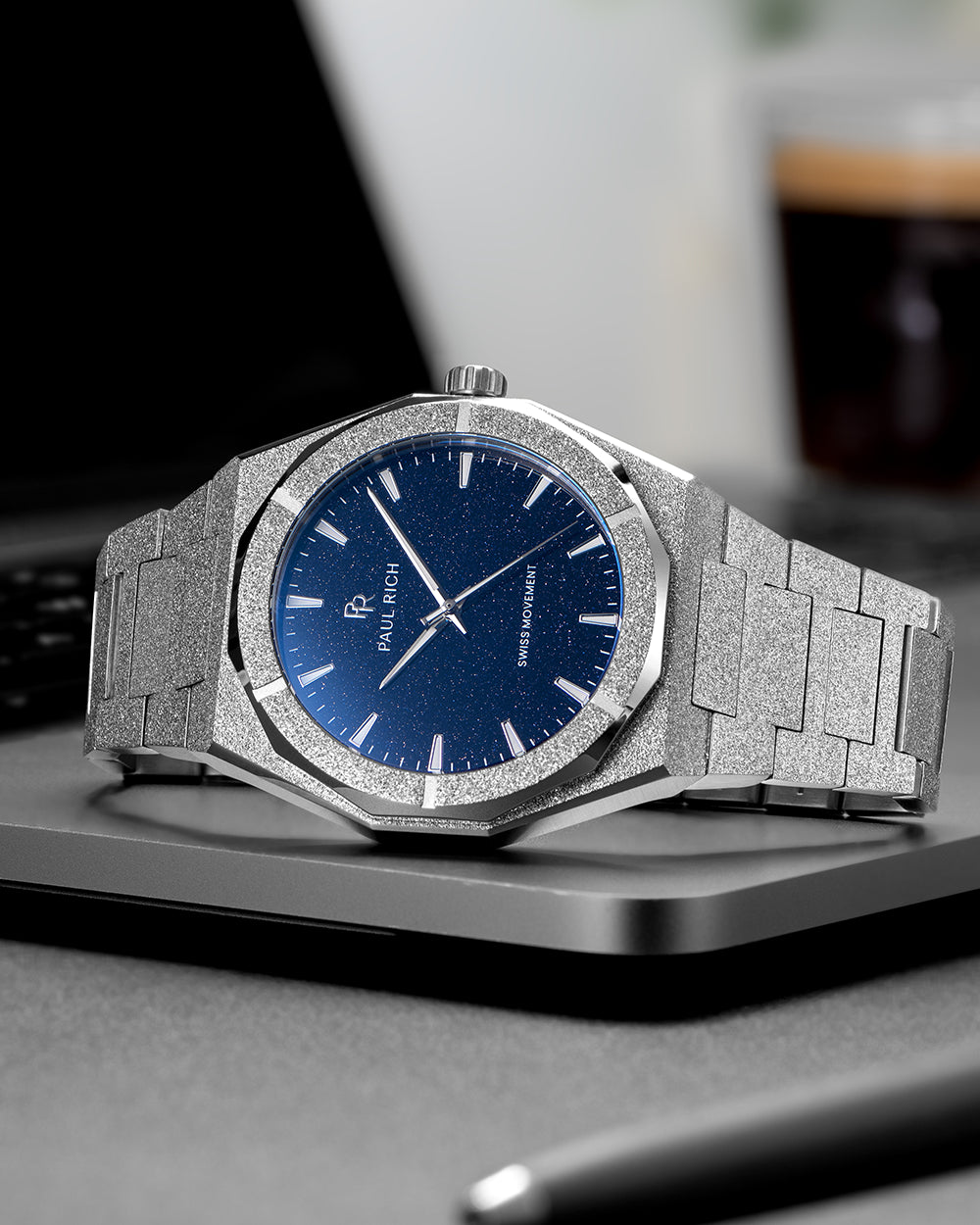 Silver frosted Paul Rich watch with blue aventurine dial resting on a closed laptop, with a blurred coffee cup in the background. Silver frosted Paul Rich watch with blue aventurine dial resting on a closed laptop, with a blurred coffee cup in the background.