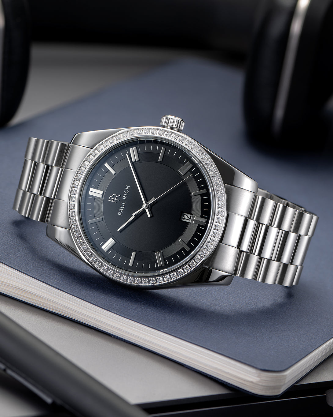 Silver-tone Paul Rich watch with black sunray dial, diamond-set bezel, and silver link bracelet, displayed on top of stacked hardcover notebooks with a pen and headphones nearby. Silver-tone Paul Rich watch with black sunray dial, diamond-set bezel, and silver link bracelet, displayed on top of stacked hardcover notebooks with a pen and headphones nearby.
