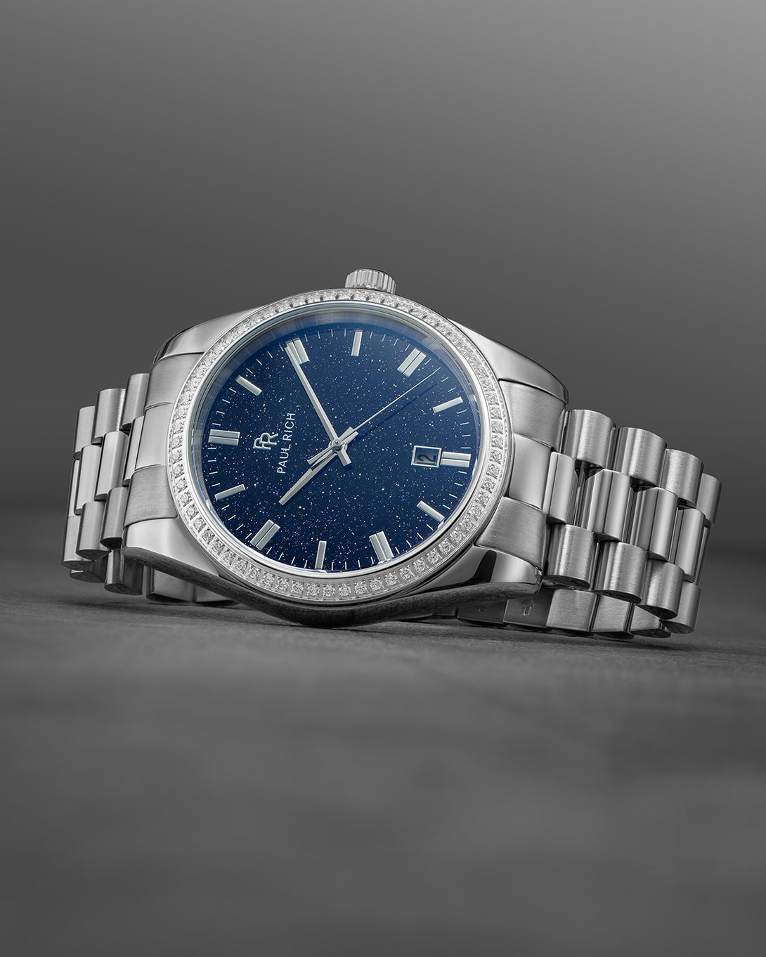 Silver wristwatch with a blue speckled dial, crystal-set bezel, and metal bracelet displayed on a dark gray surface with a gradient background. Silver wristwatch with a blue speckled dial, crystal-set bezel, and metal bracelet displayed on a dark gray surface with a gradient background.