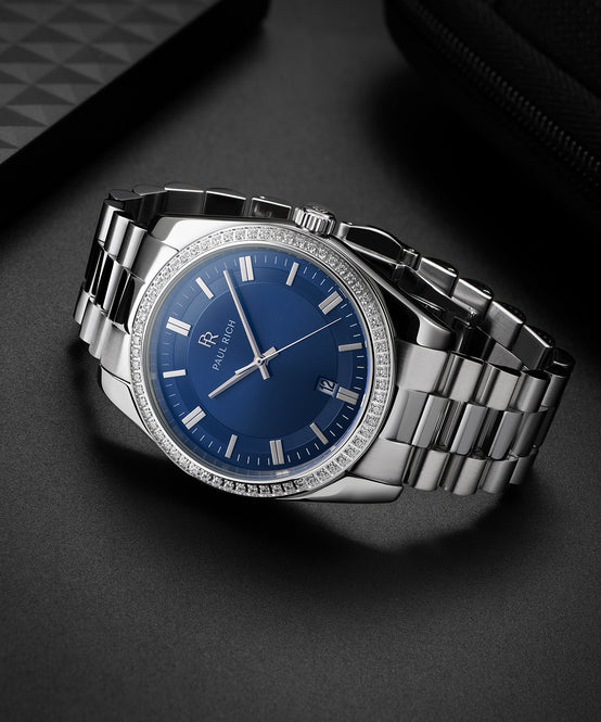Silver-tone Paul Rich watch with blue sunray dial, diamond-set bezel, and silver link bracelet, displayed on a dark matte surface beside textured black accessories. Silver-tone Paul Rich watch with blue sunray dial, diamond-set bezel, and silver link bracelet, displayed on a dark matte surface beside textured black accessories.