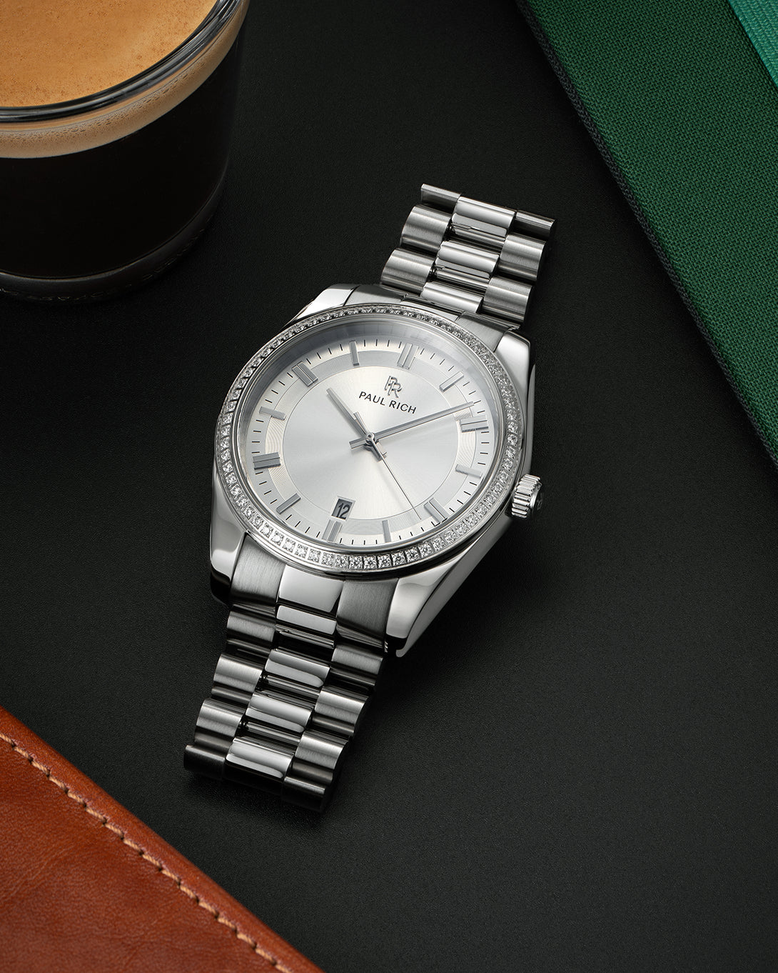 Silver-tone Paul Rich watch with silver sunray dial, diamond-set bezel, and polished silver link bracelet, displayed on a black surface with a cup of espresso, green notebook, and brown leather item. Silver-tone Paul Rich watch with silver sunray dial, diamond-set bezel, and polished silver link bracelet, displayed on a black surface with a cup of espresso, green notebook, and brown leather item.