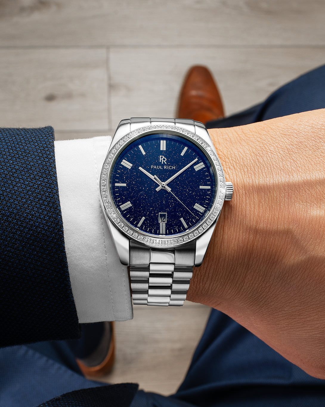 Close-up of a silver wristwatch with a blue speckled dial, worn on a man's wrist in a navy suit and brown shoes, sitting indoors. Close-up of a silver wristwatch with a blue speckled dial, worn on a man's wrist in a navy suit and brown shoes, sitting indoors.