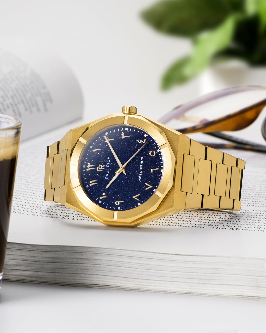 Gold watch with a blue aventurine dial and Arabic numerals, resting on an open book beside sunglasses, espresso, and a plant. Gold watch with a blue aventurine dial and Arabic numerals, resting on an open book beside sunglasses, espresso, and a plant.