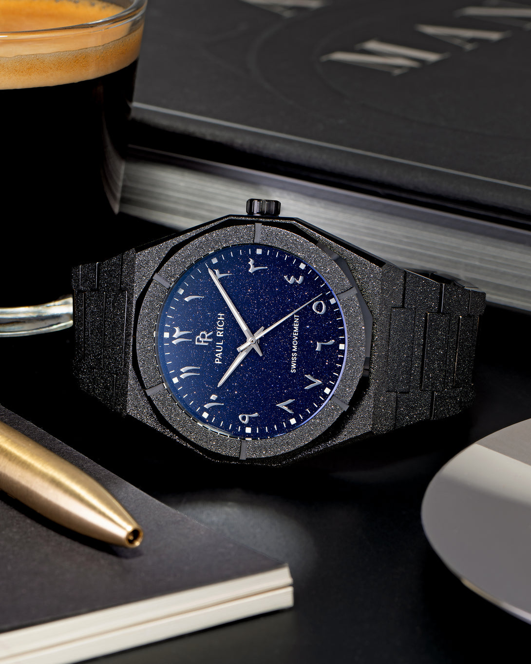 Frosted black watch with a blue aventurine dial and Arabic numerals, placed on a black desk next to a glass of espresso, gold pen, and notebook. Frosted black watch with a blue aventurine dial and Arabic numerals, placed on a black desk next to a glass of espresso, gold pen, and notebook.
