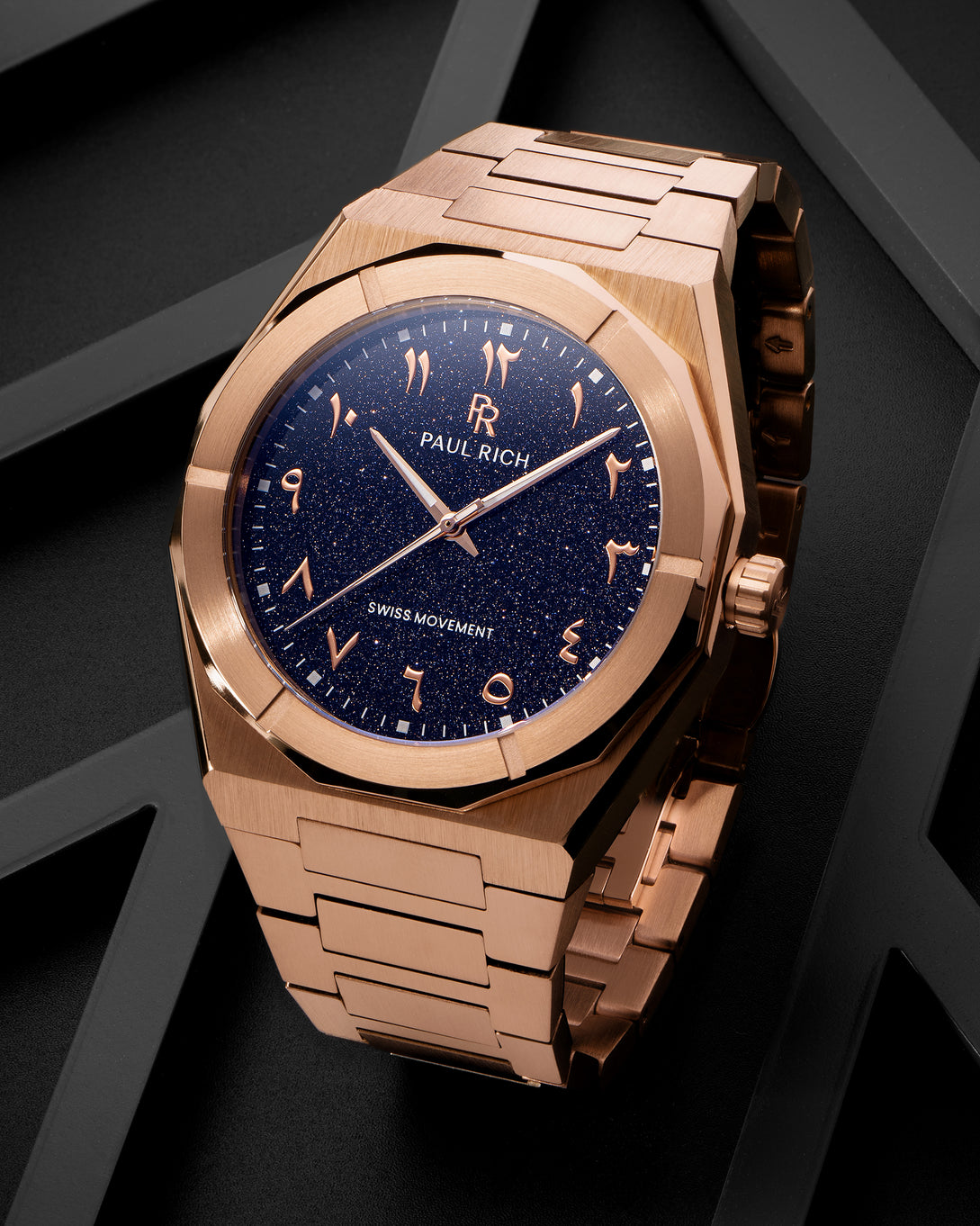 Paul Rich watch with brushed rose gold case and blue aventurine dial with Arabic numerals, displayed on a matte black geometric surface. Paul Rich watch with brushed rose gold case and blue aventurine dial with Arabic numerals, displayed on a matte black geometric surface.