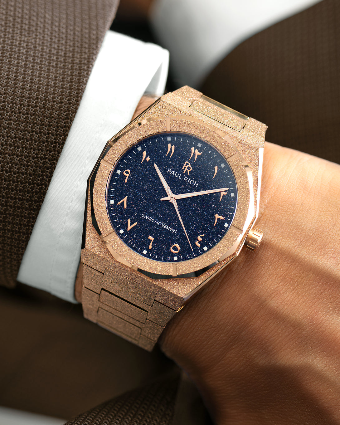 Frosted rose gold watch with blue aventurine dial and Arabic numerals, worn with a white cuff and brown textured suit. Frosted rose gold watch with blue aventurine dial and Arabic numerals, worn with a white cuff and brown textured suit.