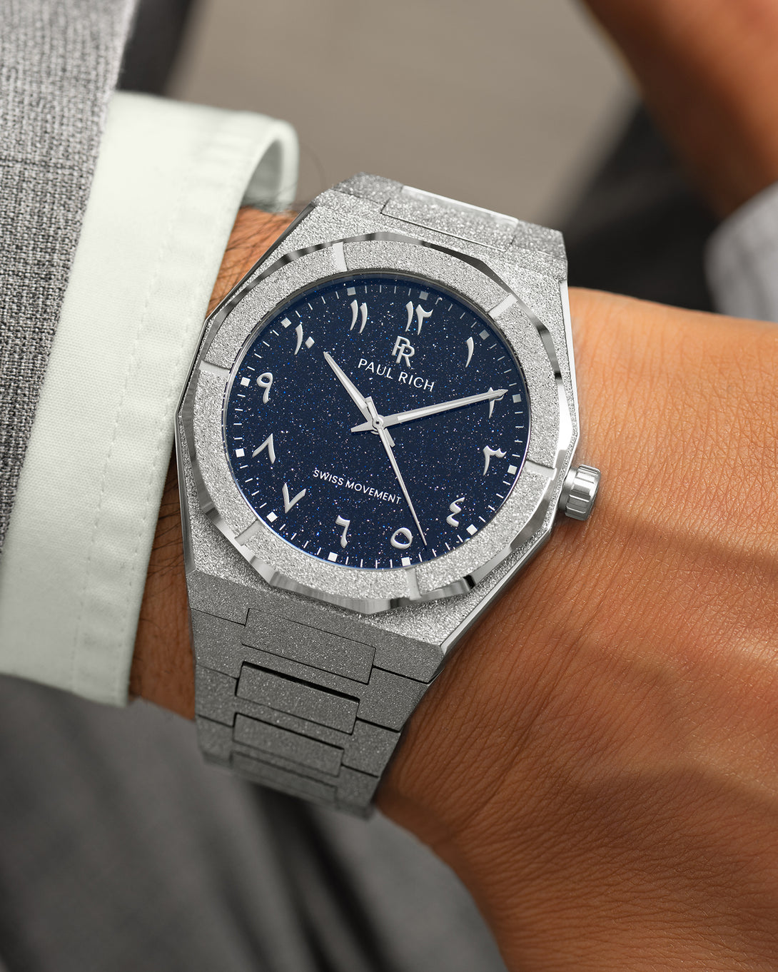 Frosted silver watch with a blue aventurine dial and Arabic numerals, worn with a light green cuff and grey suit. Frosted silver watch with a blue aventurine dial and Arabic numerals, worn with a light green cuff and grey suit.