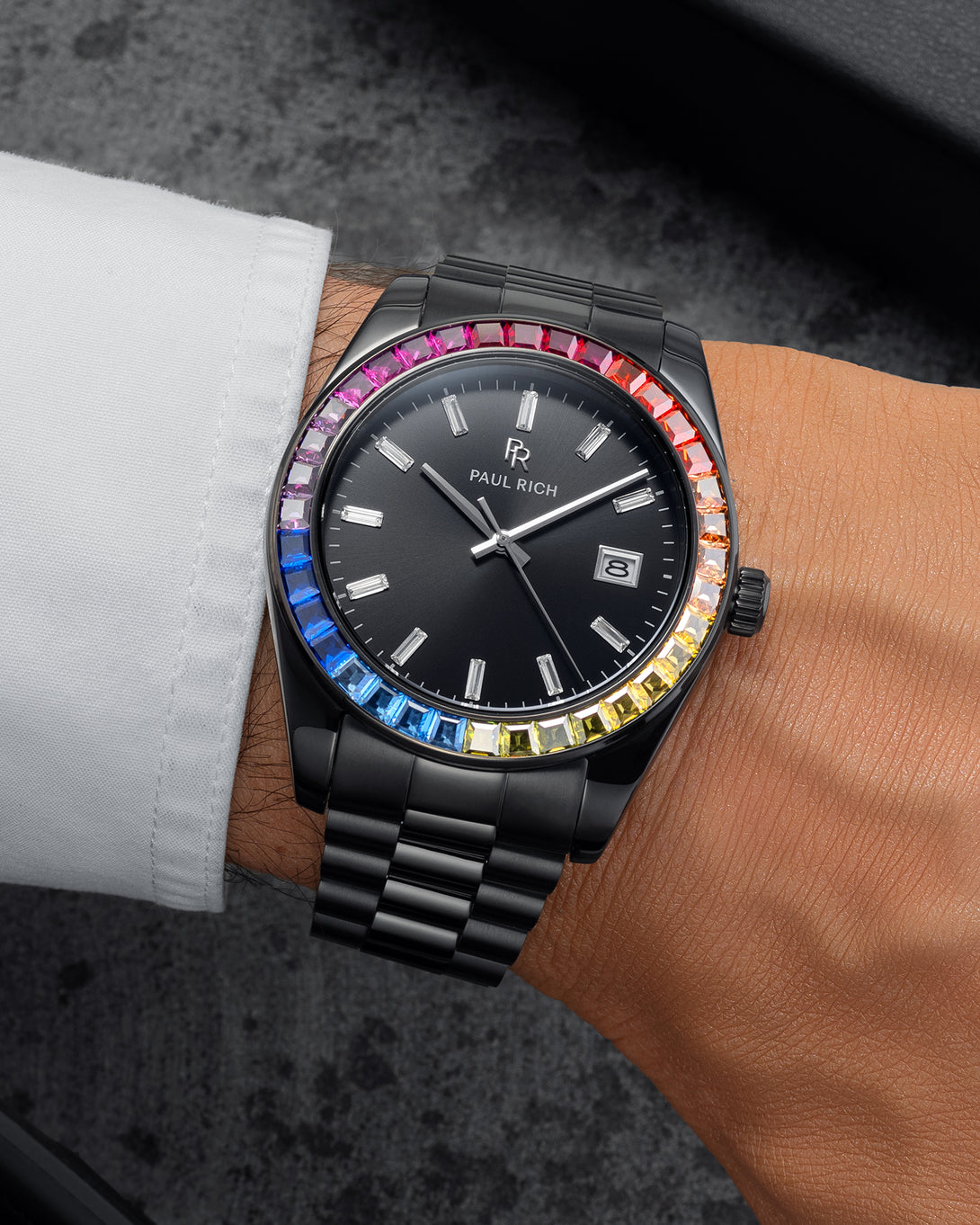 Black wristwatch with a dark dial, baguette hour markers, rainbow bezel, and black metal bracelet worn on a wrist with a white shirt sleeve. Black wristwatch with a dark dial, baguette hour markers, rainbow bezel, and black metal bracelet worn on a wrist with a white shirt sleeve.