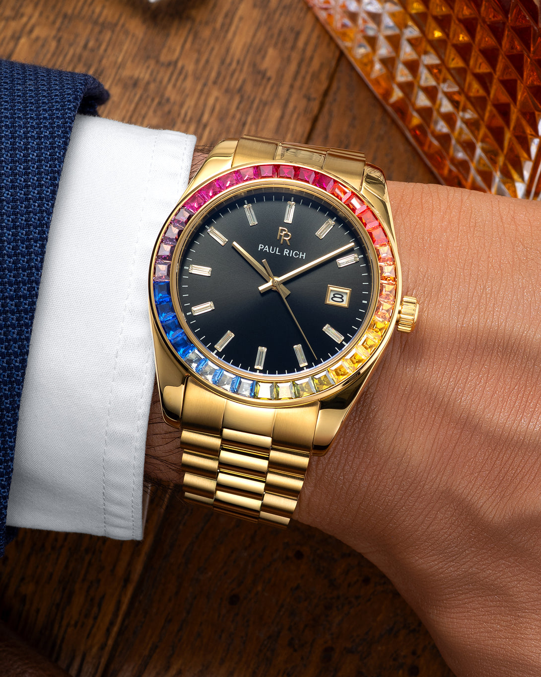 Gold wristwatch with a black dial and rainbow bezel worn on a wrist with a blue suit and white shirt, resting on a wooden surface near a glass decanter. Gold wristwatch with a black dial and rainbow bezel worn on a wrist with a blue suit and white shirt, resting on a wooden surface near a glass decanter.