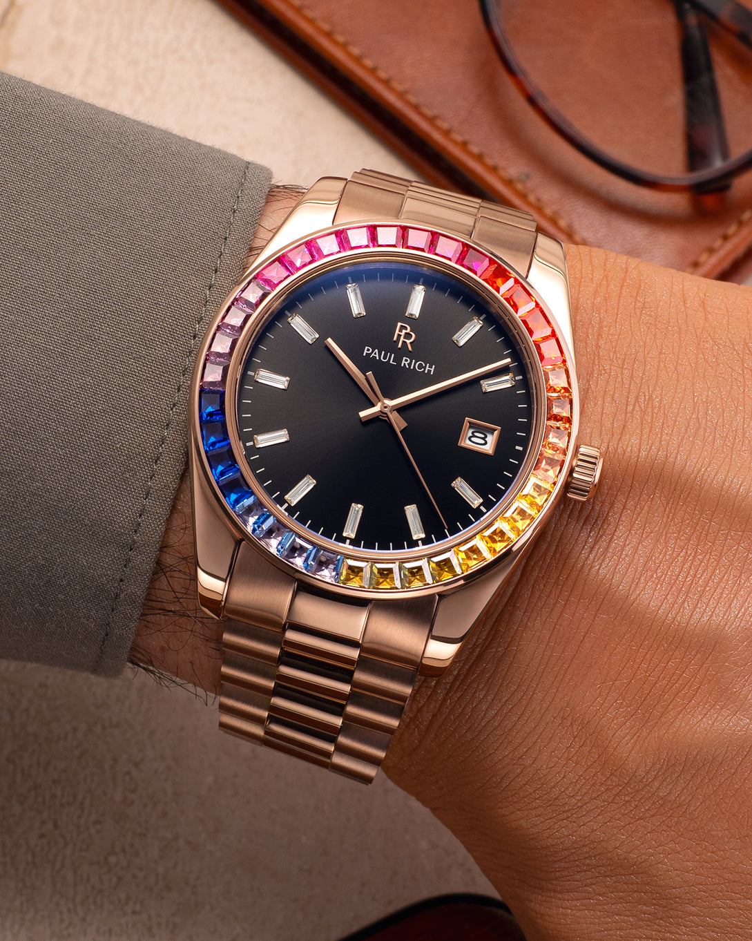 Rose gold wristwatch with a black dial and rainbow bezel worn on a wrist with a green shirt sleeve, next to a brown leather case and glasses. Rose gold wristwatch with a black dial and rainbow bezel worn on a wrist with a green shirt sleeve, next to a brown leather case and glasses.