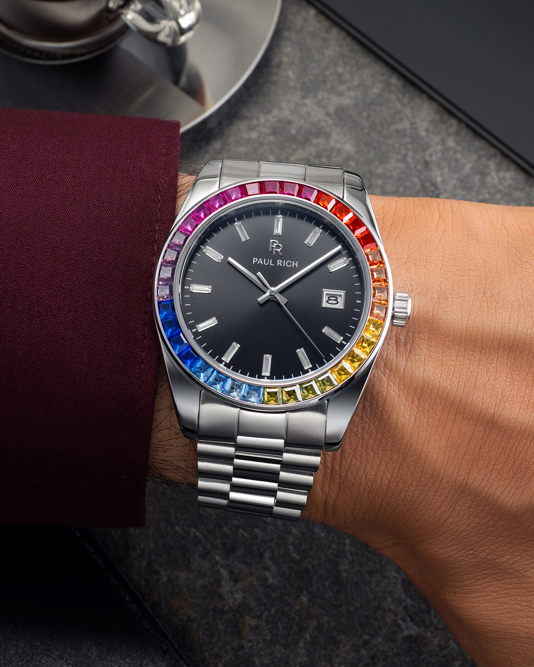 Silver wristwatch with a black dial and rainbow bezel worn on a wrist with a burgundy shirt sleeve, resting on a dark stone surface near a metal cup. Silver wristwatch with a black dial and rainbow bezel worn on a wrist with a burgundy shirt sleeve, resting on a dark stone surface near a metal cup.