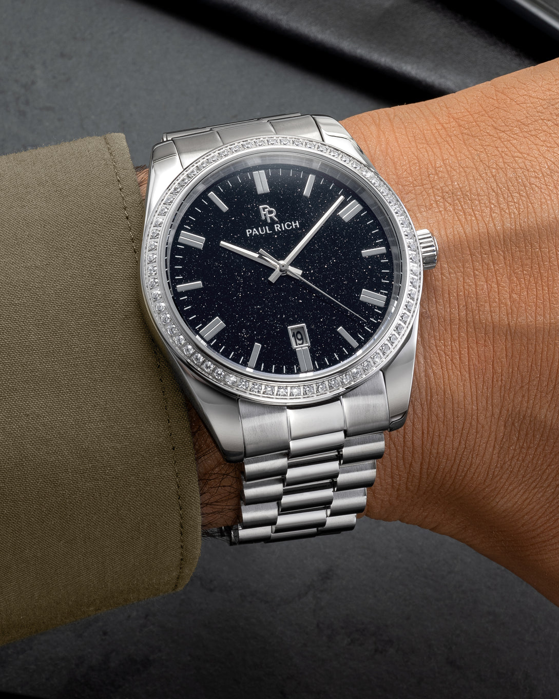 Silver wristwatch with a black aventurine dial, crystal-set bezel, and metal bracelet worn on a wrist with an olive green sleeve on a dark surface. Silver wristwatch with a black aventurine dial, crystal-set bezel, and metal bracelet worn on a wrist with an olive green sleeve on a dark surface.