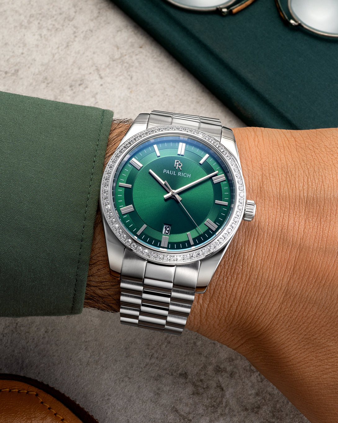 Silver wristwatch with a green sunburst dial, crystal-set bezel, and metal bracelet worn on a wrist with a green shirt sleeve. Silver wristwatch with a green sunburst dial, crystal-set bezel, and metal bracelet worn on a wrist with a green shirt sleeve.