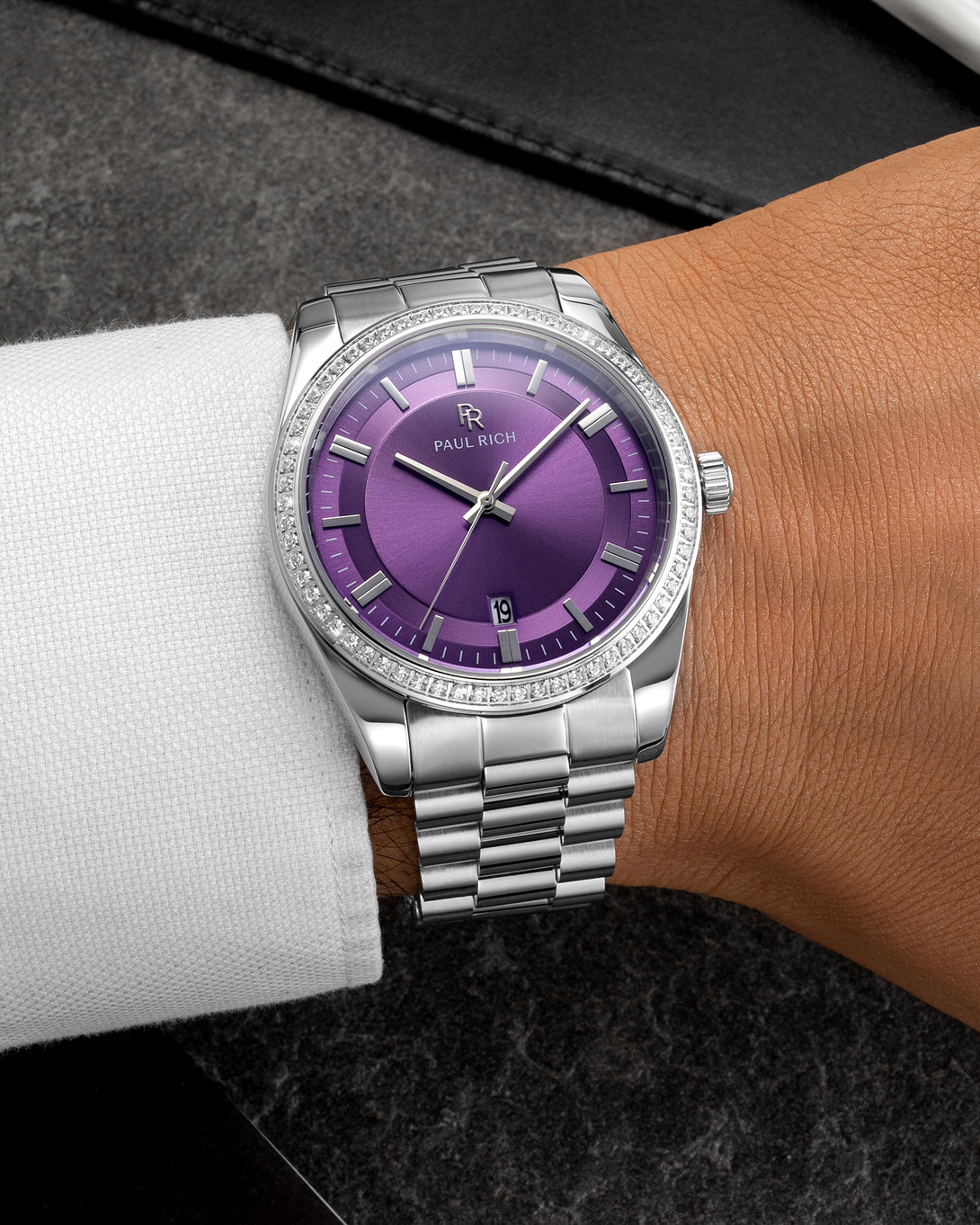Silver wristwatch with a purple sunburst dial, crystal-set bezel, and metal bracelet worn on a wrist with a white shirt sleeve over a dark surface. Silver wristwatch with a purple sunburst dial, crystal-set bezel, and metal bracelet worn on a wrist with a white shirt sleeve over a dark surface.