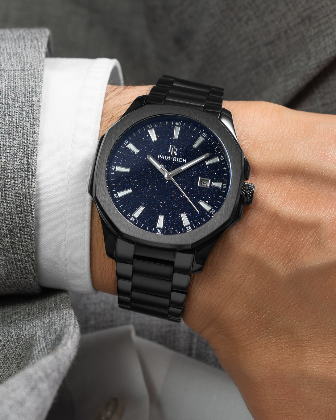Black Paul Rich watch with blue aventurine dial and date window, worn on wrist with white cuff and light grey textured suit. Black Paul Rich watch with blue aventurine dial and date window, worn on wrist with white cuff and light grey textured suit.