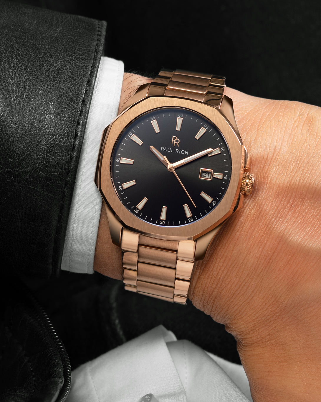 Rose gold Paul Rich watch with black sunray dial, steel bracelet, and date window, shown on wrist with white shirt and black leather jacket. Rose gold Paul Rich watch with black sunray dial, steel bracelet, and date window, shown on wrist with white shirt and black leather jacket.