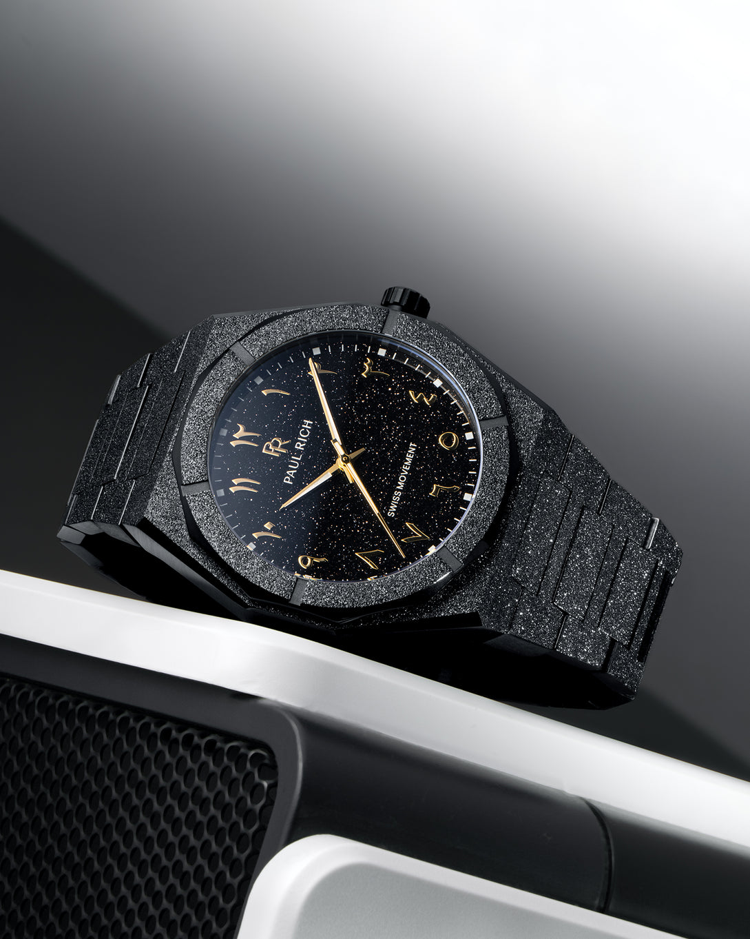 Paul Rich frosted black watch with black aventurine dial and gold Arabic numerals, resting on a silver and black speaker. Paul Rich frosted black watch with black aventurine dial and gold Arabic numerals, resting on a silver and black speaker.
