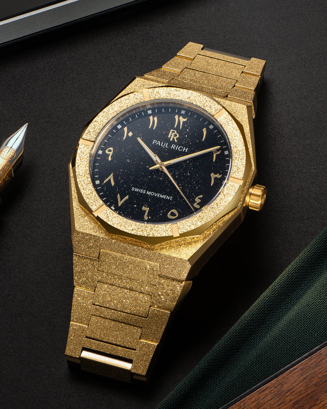 Paul Rich frosted gold watch with black aventurine dial and gold Arabic numerals, displayed on a black surface near a fountain pen. Paul Rich frosted gold watch with black aventurine dial and gold Arabic numerals, displayed on a black surface near a fountain pen.