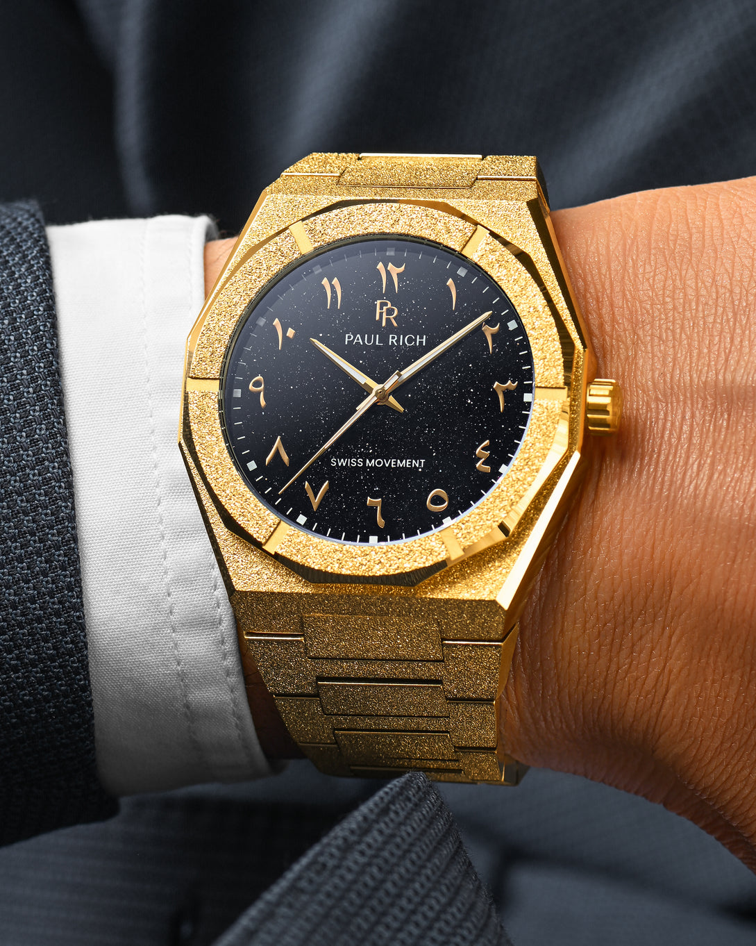 Close-up of Paul Rich frosted gold watch with Arabic numerals and black aventurine dial, worn on wrist with dark textured suit. Close-up of Paul Rich frosted gold watch with Arabic numerals and black aventurine dial, worn on wrist with dark textured suit.