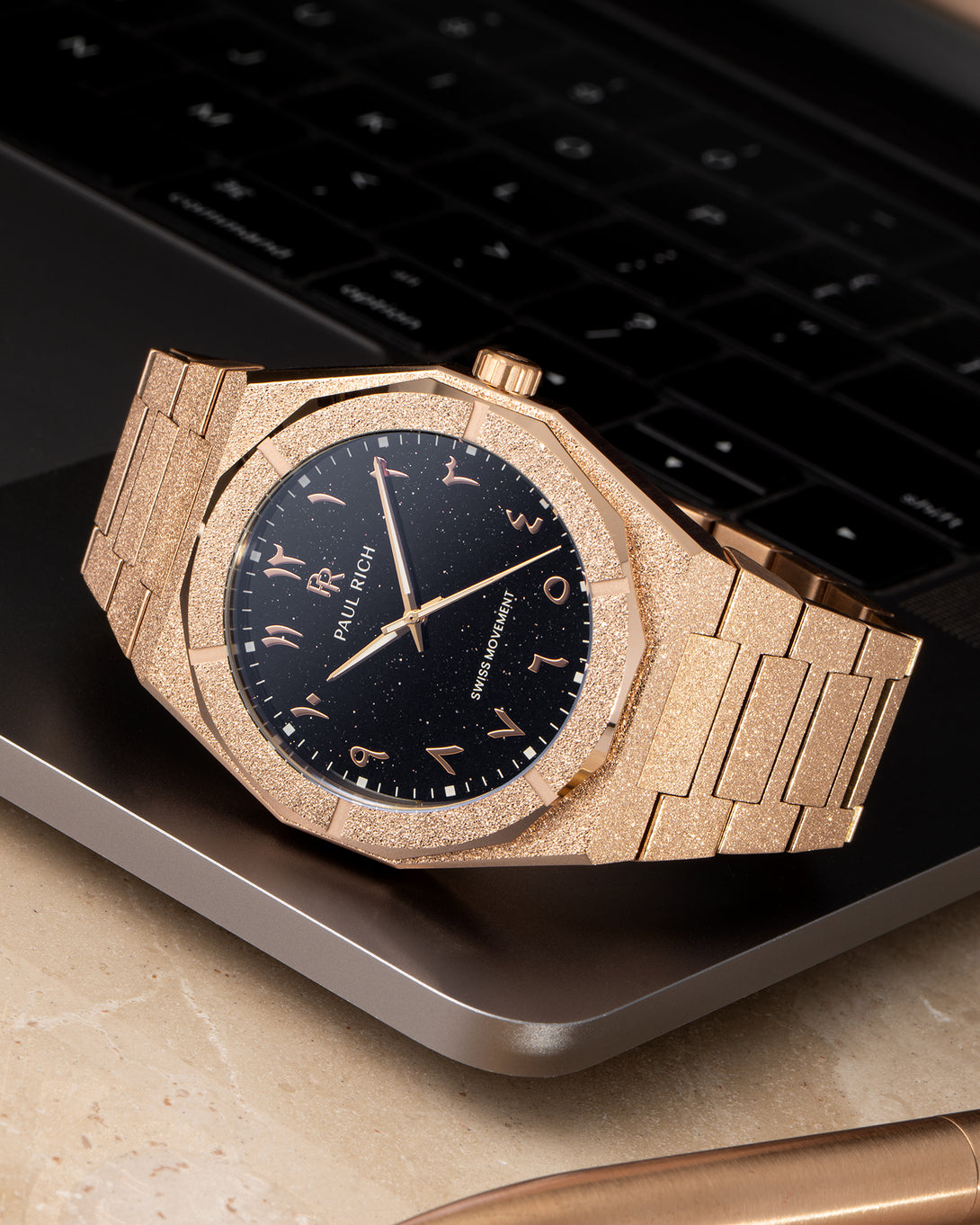 Paul Rich frosted rose gold watch with black aventurine dial and rose gold Arabic numerals, resting on a MacBook corner. Paul Rich frosted rose gold watch with black aventurine dial and rose gold Arabic numerals, resting on a MacBook corner.