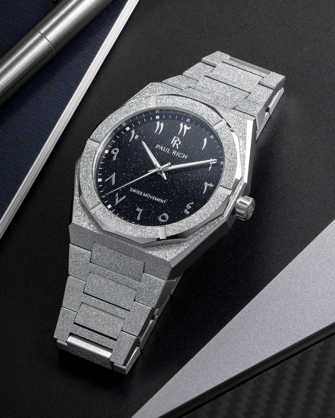 Paul Rich frosted silver watch with black aventurine dial and silver Arabic numerals, displayed on a black surface near a notebook. Paul Rich frosted silver watch with black aventurine dial and silver Arabic numerals, displayed on a black surface near a notebook.