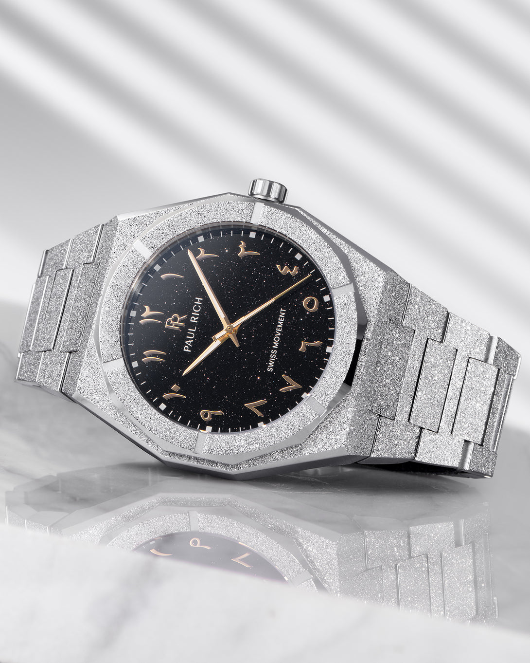Paul Rich frosted silver watch with black aventurine dial and gold Arabic numerals, resting on a reflective marble surface. Paul Rich frosted silver watch with black aventurine dial and gold Arabic numerals, resting on a reflective marble surface.