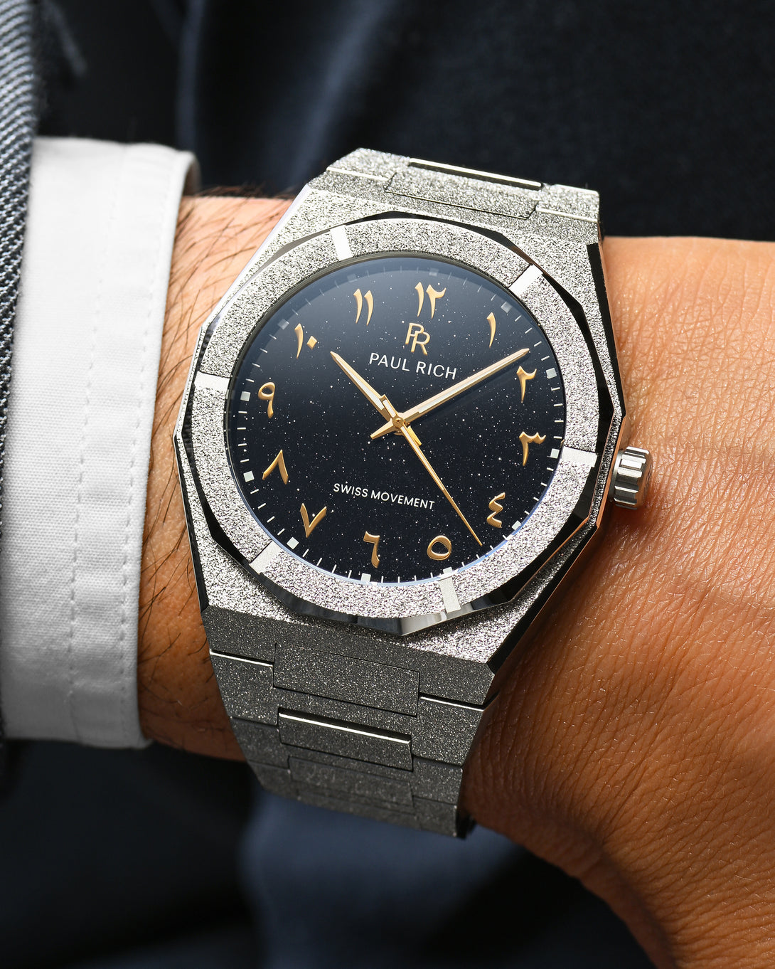 Close-up of Paul Rich frosted silver watch with gold Arabic numerals and black aventurine dial, worn with a white cuff and dark jacket. Close-up of Paul Rich frosted silver watch with gold Arabic numerals and black aventurine dial, worn with a white cuff and dark jacket.