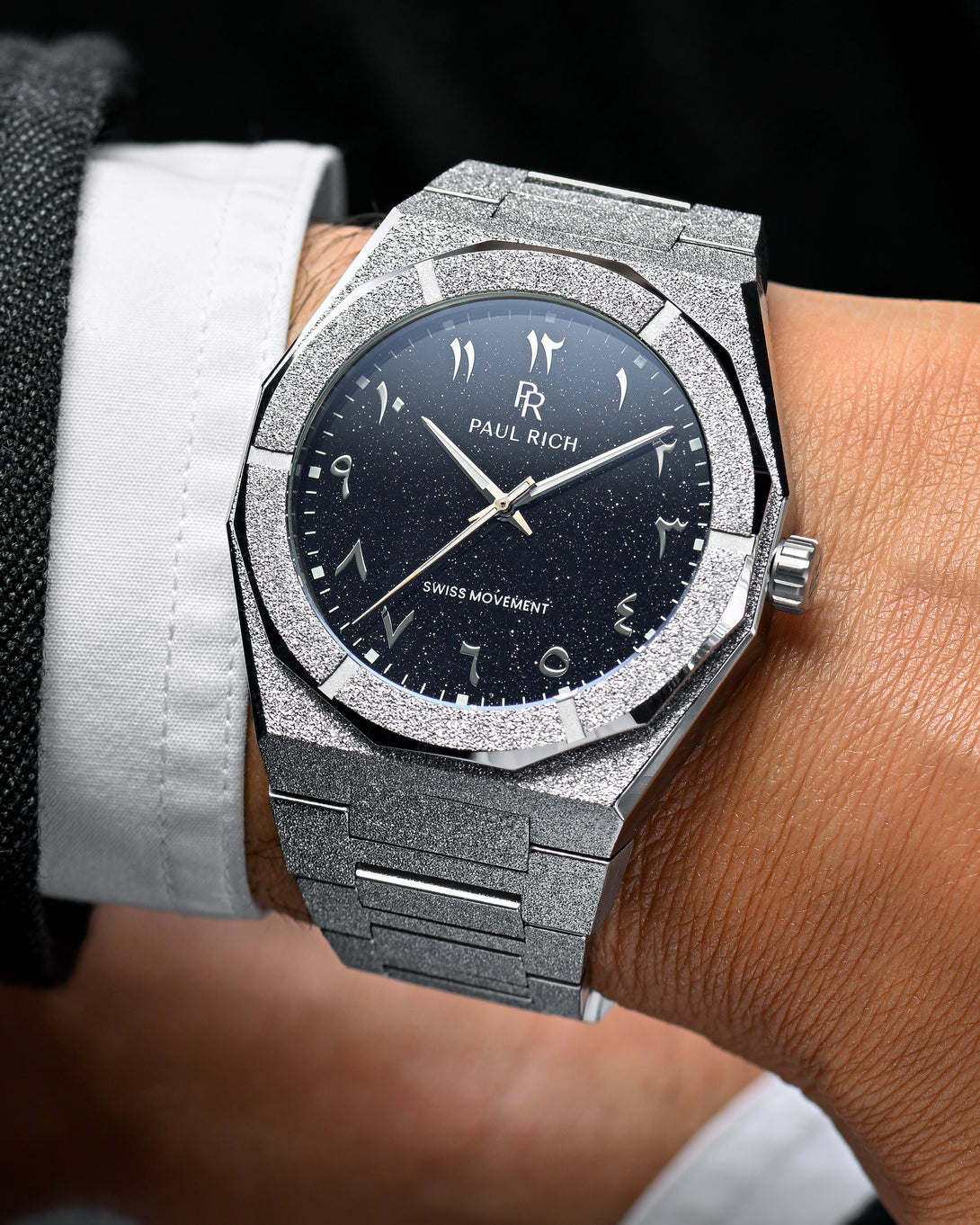Close-up of Paul Rich frosted silver watch with silver Arabic numerals and black aventurine dial, worn with white shirt and dark jacket. Close-up of Paul Rich frosted silver watch with silver Arabic numerals and black aventurine dial, worn with white shirt and dark jacket.