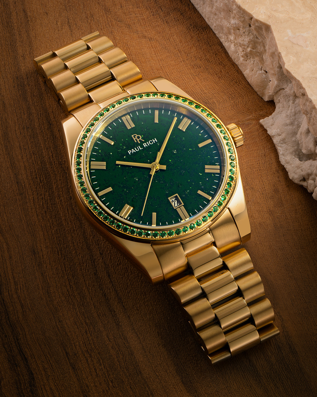 Gold watch with green aventurine dial and green gemstone-set bezel displayed on a wooden surface near a stone edge, showing the Paul Rich logo and date window. Gold watch with green aventurine dial and green gemstone-set bezel displayed on a wooden surface near a stone edge, showing the Paul Rich logo and date window.