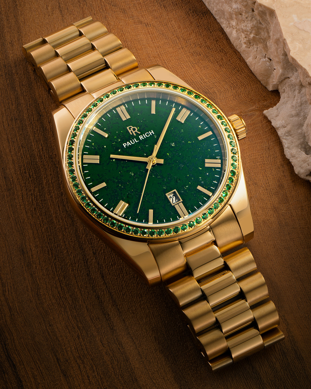 Gold watch with green aventurine dial and green gemstone-set bezel displayed on a wooden surface near a stone edge, showing the Paul Rich logo and date window. Gold watch with green aventurine dial and green gemstone-set bezel displayed on a wooden surface near a stone edge, showing the Paul Rich logo and date window.