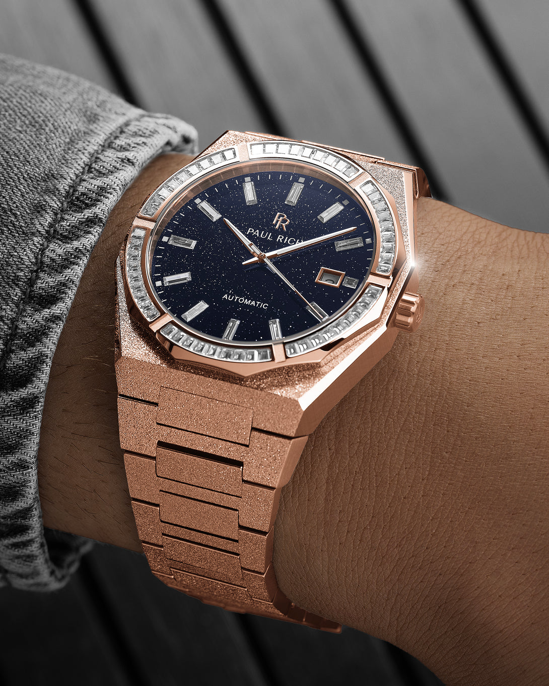 Rose gold frosted watch with a dark blue sparkle dial and baguette-cut crystal bezel on a wrist wearing a gray textured sleeve. Rose gold frosted watch with a dark blue sparkle dial and baguette-cut crystal bezel on a wrist wearing a gray textured sleeve.