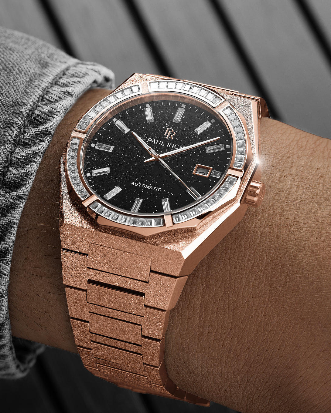 Rose gold frosted watch on a wrist with a black aventurine dial, baguette-cut stones around the bezel, and a textured rose gold bracelet. Rose gold frosted watch on a wrist with a black aventurine dial, baguette-cut stones around the bezel, and a textured rose gold bracelet.