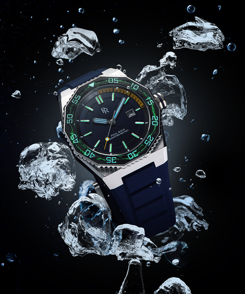 Diver-style watch with brushed steel case, black bezel, glowing green markers, blue dial, and rubber strap, shown underwater with air bubbles. Diver-style watch with brushed steel case, black bezel, glowing green markers, blue dial, and rubber strap, shown underwater with air bubbles.
