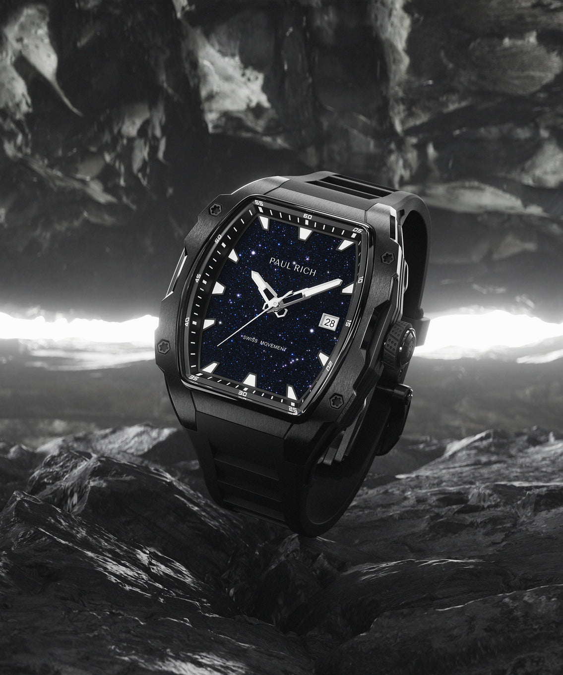 Black Paul Rich watch with a tonneau-shaped case and blue aventurine dial, featuring white markers, date window, and black rubber strap, displayed upright on rugged black rocks with a glowing cavern-like background. Black Paul Rich watch with a tonneau-shaped case and blue aventurine dial, featuring white markers, date window, and black rubber strap, displayed upright on rugged black rocks with a glowing cavern-like background.