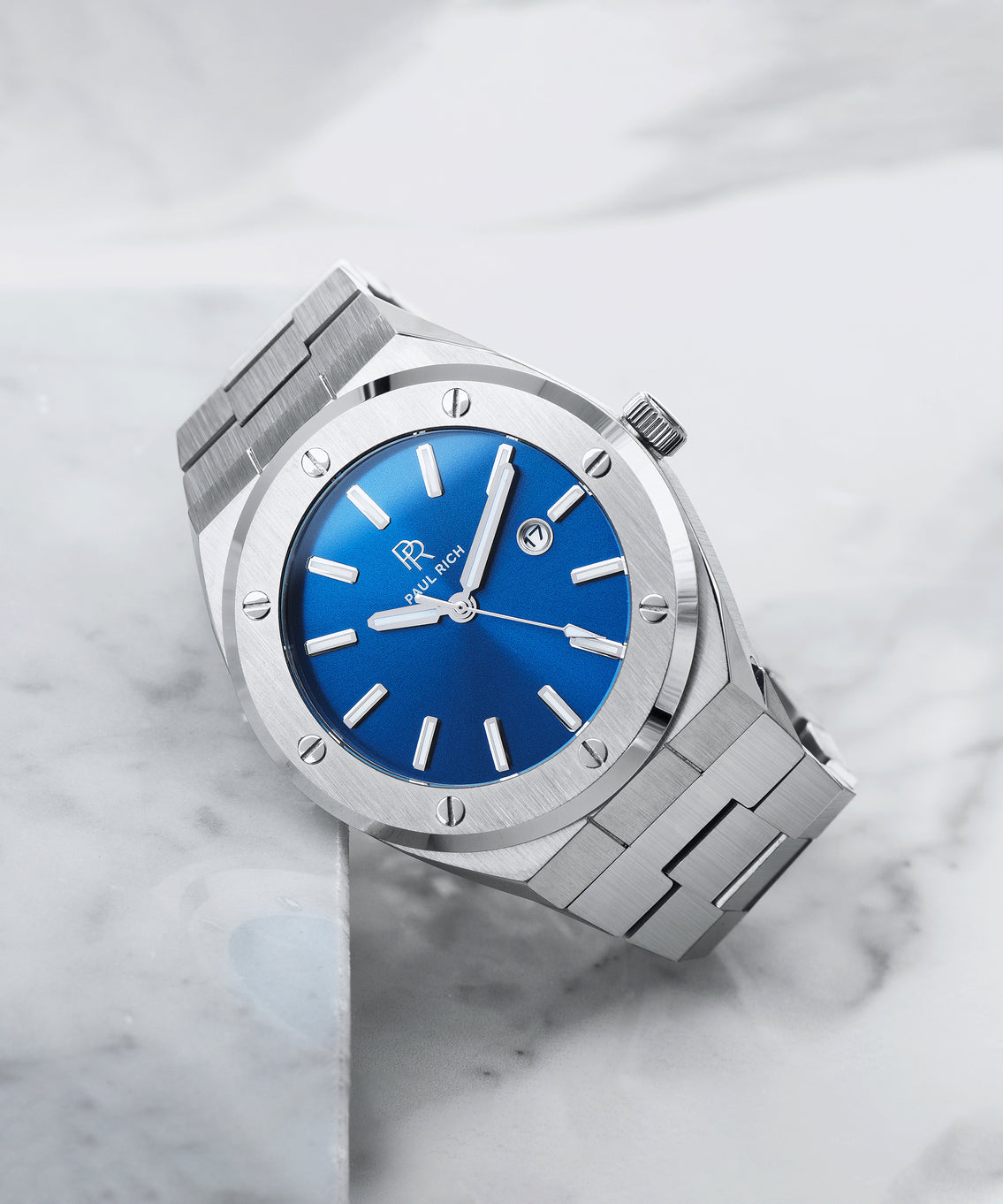 Paul Rich brushed silver watch with vivid blue sunray dial and date window, placed diagonally on white marble surface. Paul Rich brushed silver watch with vivid blue sunray dial and date window, placed diagonally on white marble surface.