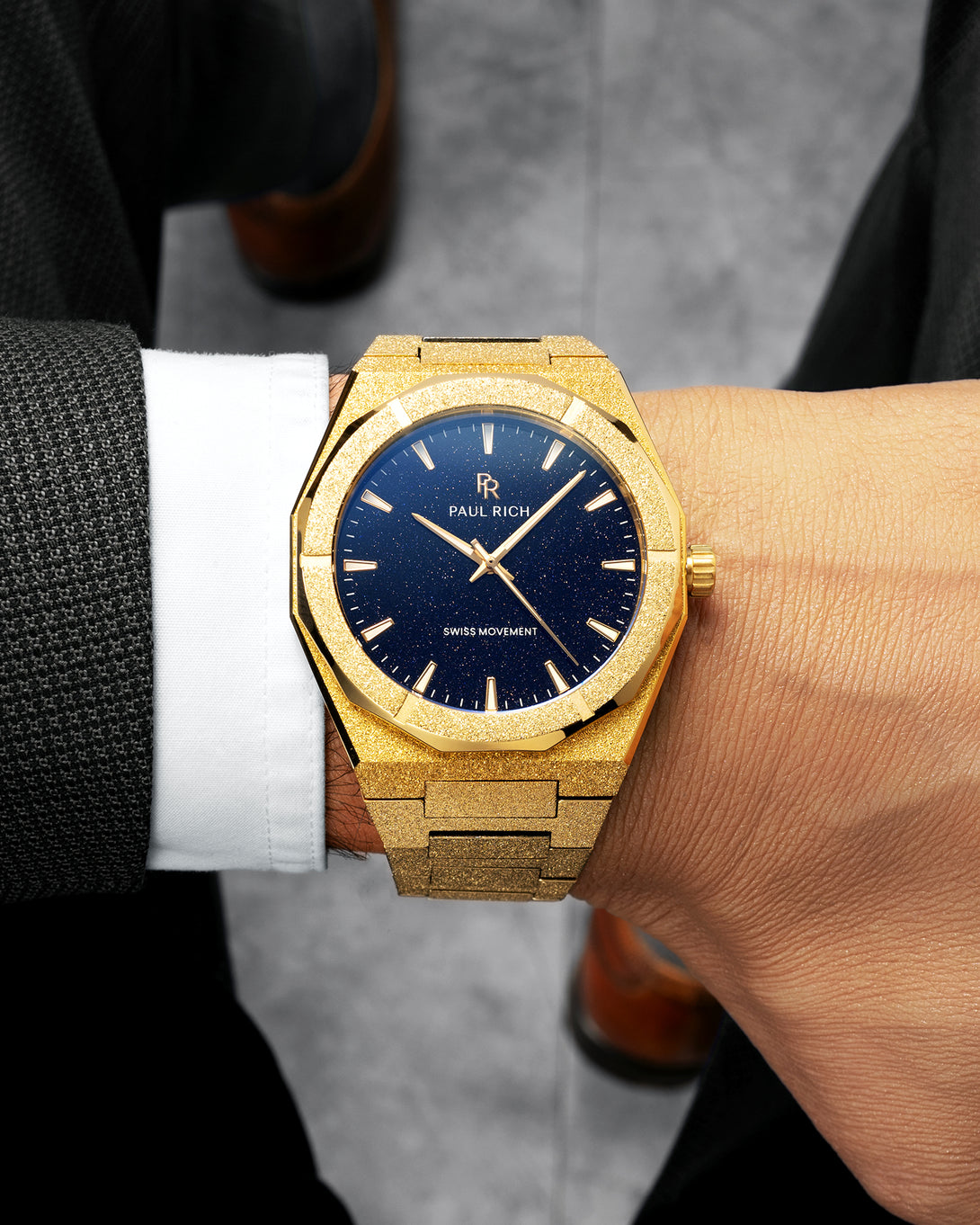 Gold frosted Paul Rich watch with blue aventurine dial worn on wrist, paired with a dark suit, white cuff, and brown shoes. Gold frosted Paul Rich watch with blue aventurine dial worn on wrist, paired with a dark suit, white cuff, and brown shoes.