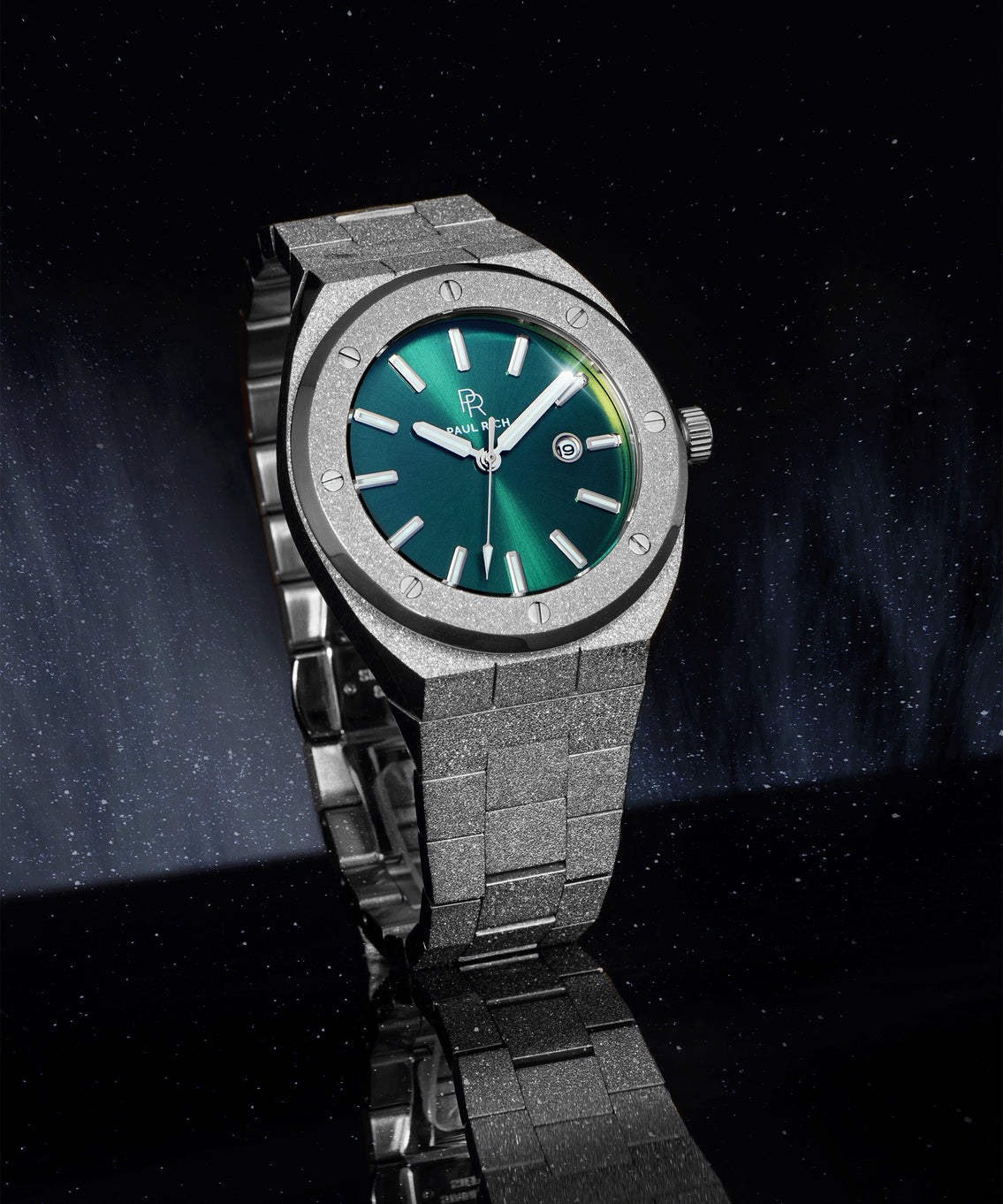 Paul Rich frosted watch with green sunray dial, silver markers, and date window, shown at an angle on a reflective black surface. Paul Rich frosted watch with green sunray dial, silver markers, and date window, shown at an angle on a reflective black surface.