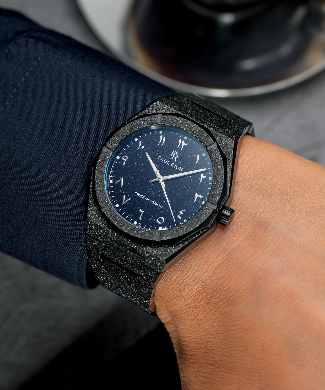 Frosted black watch with a blue aventurine dial and Arabic numerals, worn with a navy cuff, next to a glass cup of espresso. Frosted black watch with a blue aventurine dial and Arabic numerals, worn with a navy cuff, next to a glass cup of espresso.