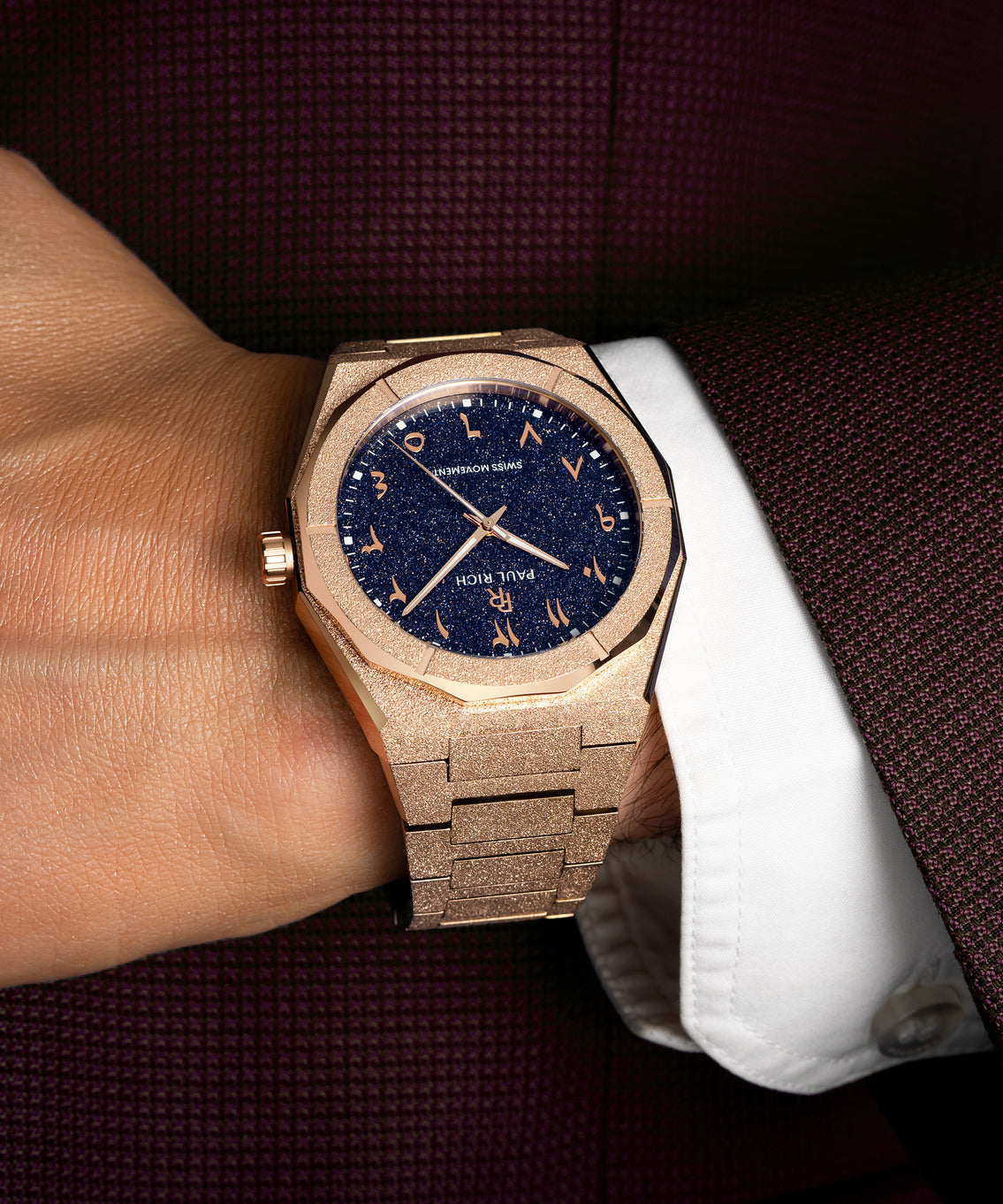 Frosted rose gold watch with blue aventurine dial and Arabic numerals, worn with a white cuff and burgundy textured suit. Frosted rose gold watch with blue aventurine dial and Arabic numerals, worn with a white cuff and burgundy textured suit.