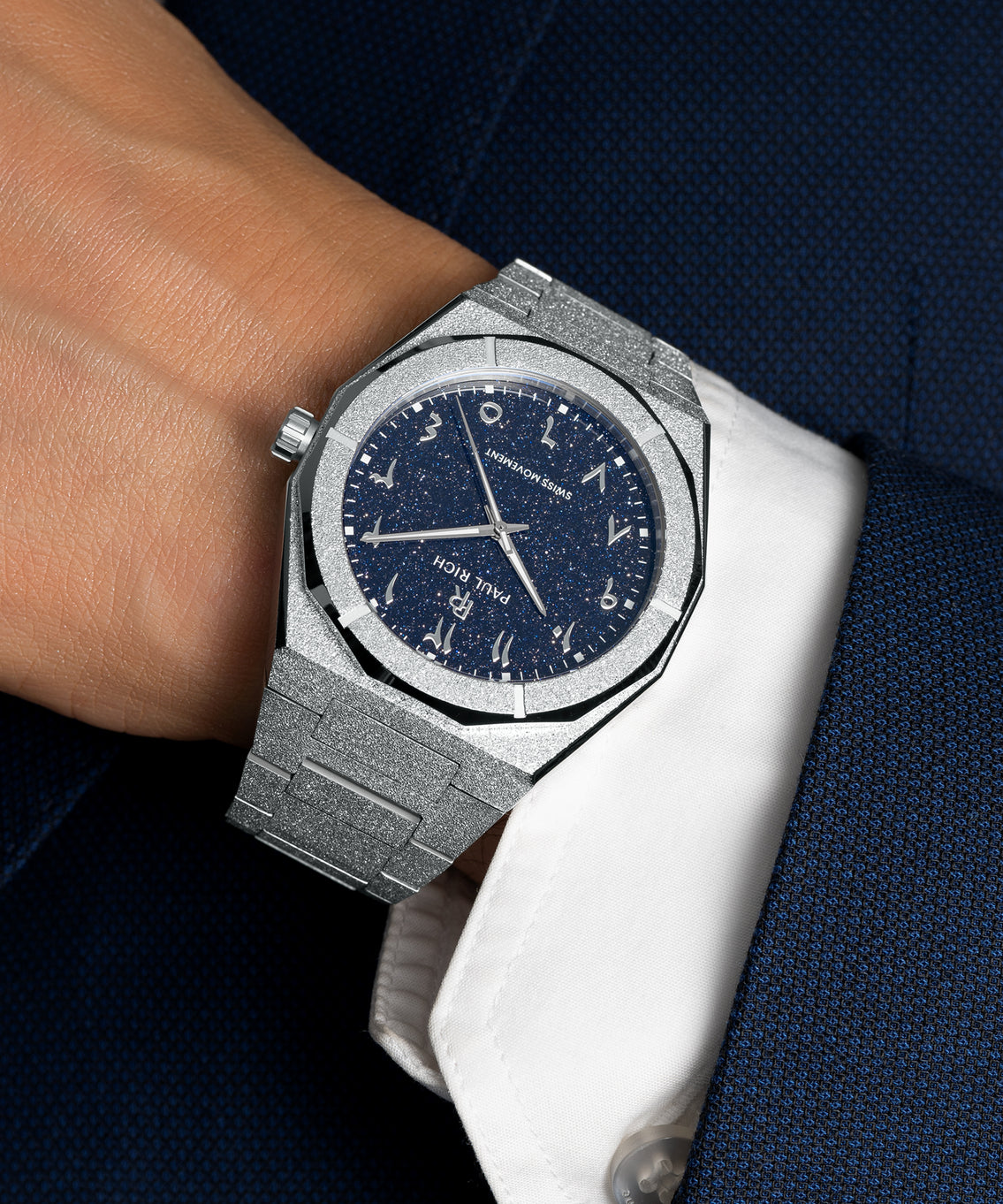 Frosted silver watch with a blue aventurine dial and Arabic numerals, worn with a white cuff and dark blue textured suit. Frosted silver watch with a blue aventurine dial and Arabic numerals, worn with a white cuff and dark blue textured suit.